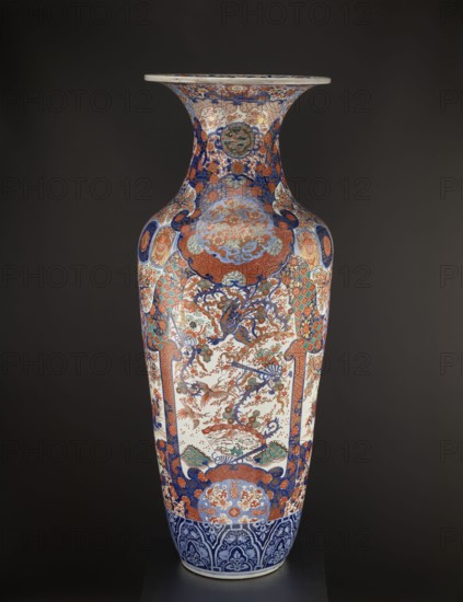Vase, Fujisaki, Tahei,  craftsman, Japanese art, Japanese culture, Meiji 1868-1912, vases, birds, floral decorations, plant ornaments, cherries, phoenixes iconographic, ceramics, decorative vessels, colored porcelain, imari, sakura iconographic, turtles, chrysanthemums, gourds, fish, cranes, fans, dragons, butterflies, kikko pattern, kojitsunagi pattern, seigaiha pattern, hoo phoenix. Ovoid vase, slender belly turning into a long neck with a flared spout. Four-reserve decoration with a cherry blossom tree and alternating phoenixes; multicolored background with geometric-floral patterns. ca. 1870–1880 Meiji Period, 1868–1912. Japan; Hizen province, Arita. Vessel; height 122.0 cm, diameter 44.0 cm, weight 43.0 kg., A tall decorative vase with a flared rim standing against a dark background, covered in intricate painted patterns and scenes in colors including blue, orange, red, white, green, and gold; the neck and shoulders feature dense floral and geometric motifs with circular medallion...
