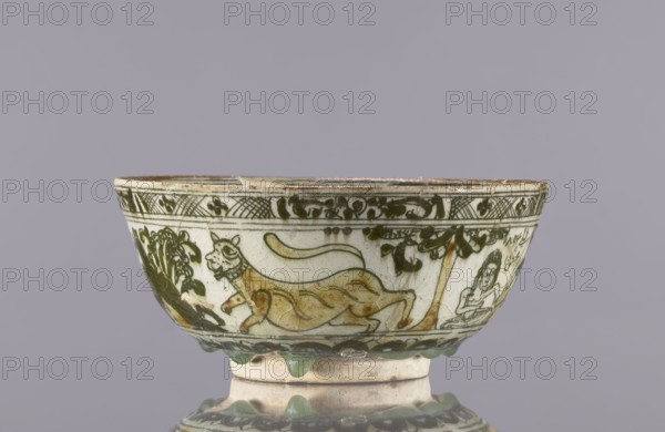 Bowl, late 19th century, Iran. Ceramic vessel tableware/utility ceramic, height 8.5 cm, diameter 19 cm. Persian Qajar dynasty style. Decoration on the rim: Rostam, with a dragon and Majnun i Lejla and the date 1893. Figural scenes, dragons, birds, literary motifs; also depicts lutes and a jambiyah., A shallow ceramic bowl with a rounded foot showing a painted narrative band around its exterior, featuring a reclining animal figure rendered in yellowish-brown with dark green outlines and a stylized tree or foliage nearby in dark green, with an adjacent human figure drawn in fine dark green lines; the upper rim area is decorated with a repeating dark green linear and foliate border, and the lower rim has a row of raised scalloped green drips above the pale off-white body; visible colors include pale off-white, dark green, yellowish-brown, and hints of brown on the rim, set against a neutral gray background.