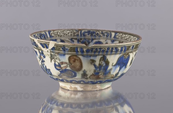 Bowl, ceramics, Persian, Qajar dynasty 1785–1925, 2nd half of the 19th century, Iran. Vessel, height 9 cm, diameter 20 cm. Decoration: gazelles, figural scenes, lions, female figures; decoration on rims: figure with nargile, other figures, lion, palmettes, architecture, inscriptions. Maker, provenance includes Abdullazad Mirza Baba fl. ante 1939., A ceramic bowl with visible chips and a repaired break sits on a reflective surface; the bowl's exterior is decorated with painted figurative scenes and ornamental bands in blue, white, brown, and black, with touches of green and beige in the glaze and repair areas. The rim shows a thin brown edge and the interior near the rim displays a decorative band in blue and black. The body of the bowl features blue floral and wave-like motifs alternating with small painted humanlike figures in blue and brown tones, and a larger blue animallike form toward one side. The foot of the bowl is unglazed or worn, showing beige and brown clay, and the glossy ...