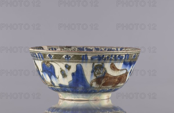 Bowl, Qajar dynasty 1785–1925, 2nd half of the 19th century, Iran; ceramics; vessel, height 9 cm, diameter 20 cm. Decoration: gazelles, figural scenes, lions, female figures; decoration on rims: figure with nargile, other figures, lion, palmettes, architecture; inscriptions. Culture: Persian Islamic art., A shallow ceramic bowl sits on a reflective surface, showing a rounded foot and slightly flared rim; the bowl is decorated with painted motifs including a stylized animal figure and flowing organic shapes. The visible colors include blue, brown, green, white, and hints of beige. The rim and upper band feature patterned ornamentation, while the main body shows a mix of blue washes and brown-painted details forming the animal and surrounding shapes. The glaze has a glossy finish with subtle crackle and areas of wear or discoloration near the base.