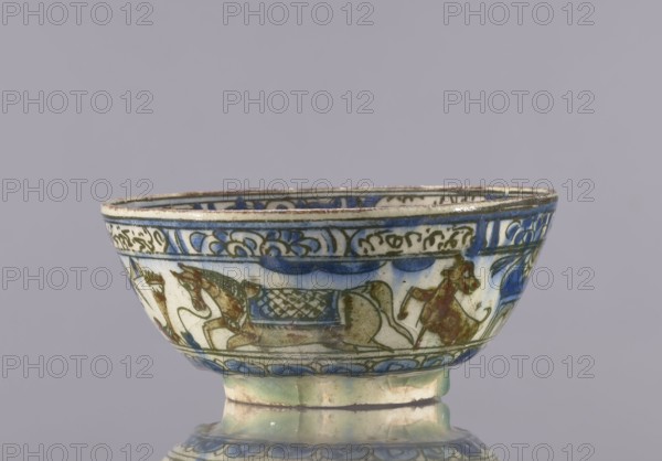 Bowl, 2nd half of the 19th century, Iran Qajar dynasty, Persian; glazed ceramic vessel tableware, height 7.5 cm, diameter 17.4 cm. Decoration: knightly and figurative scenes with horses and falcons; rims decorated with figures, architecture, flowers, animals, palmettes, and inscriptions. Cultural/ stylistic: Persian, Islamic art., A shallow ceramic bowl with a rounded foot, shown from the side, decorated with painted figurative scenes and ornamental bands; the bowl displays a palette of blue, green, brown, cream, and pale turquoise, with darker blue outlining and accents, and areas of brownish-red on the painted figures; the rim shows a thin darker edge and the interior and exterior carry repeating floral and scroll motifs, while the exterior band includes stylized animal-like figures and a patterned rectangular panel, all rendered in the same limited color range against a cream background, set against a plain light gray-purple backdrop.