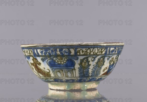 Bowl, 2nd half of the 19th century, Iran. Ceramic glazed tableware vessel height 7.5 cm; diameter 17.4 cm. Qajar dynasty 1785–1925. Decoration: knightly and figurative scenes with horses and falcons; rims decorated with figures, architecture, flowers, animals, palmettes, and inscriptions. Persian, Islamic art; ceramics., A low ceramic bowl with decorative painted designs sits on a reflective surface against a plain gray background; the bowl has a rounded body and a short foot with glaze drips visible at the base, and its exterior shows intricate painted motifs in blue, green, brown, and cream tones including floral shapes and stylized patterns, with areas of worn or aged glazing and a thin darker rim along the top edge.