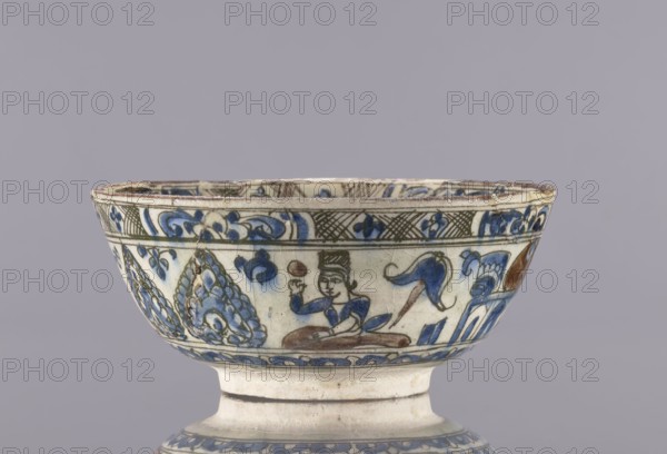 Bowl, Abdullazad Mirza Baba fl. ante 1939, Islamic art, Persian, Qajar dynasty 1785–1925; 2nd half of the 19th century, Iran. Glazed ceramic household vessel/tableware bowl; height 8 cm, diameter 19 cm. Decoration: figurative scenes including female images, dragons, demons, figures, and a horse; palmettes; rim decoration resembling architecture ziggurat., A shallow ceramic bowl on a short foot with painted decoration shows a scene of a seated human figure wearing a hat and holding a round object, surrounded by stylized plant motifs and abstract flourishes; the painted palette includes blue, white, brown, green, and cream tones; the bowl rim has a decorative band and the surface shows signs of age and wear.
