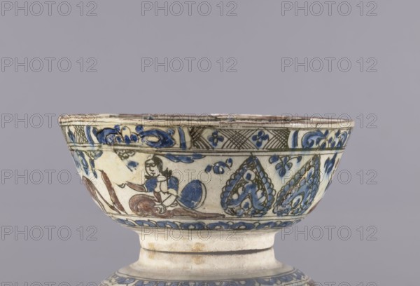 Bowl, Abdullazad Mirza Baba fl. ante 1939, Islamic art, Persian, Qajar dynasty 1785–1925, 2nd half of the 19th century, Iran. Glazed ceramic household vessel tableware, bowl; height 8 cm, diameter 19 cm. Decoration: figurative scenes with female images, dragons, demons, figures, horse, palmettes; rim decoration resembling architecture ziggurat., A ceramic bowl with a short pedestal base shown against a gray background, decorated with hand-painted motifs in blue, dark green, brown, and cream. The bowl's exterior band features floral and leaf-like patterns, curving scrolls, and a scene with a seated human figure rendered in blue and brown tones. The rim area has a narrow decorative border with intersecting hatch patterns and small floral elements in blue and green. The lower edge of the bowl above the foot has a repeating wave or petal border painted in blue. The surface shows areas of glaze variation and slight wear along the rim and foot.