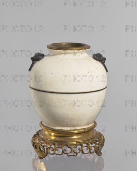 Gilded bronze mounted vase - lamp body, Chinese art, Chinese culture, Qing 1644–1911, decorative vessel, vase/lamp, cracklework decoration, 18th century, China, vessel, height 33 cm, A rounded ceramic vessel with a pale cream surface showing a network of fine crackle and some small darker speckles, featuring a short neck with a thin dark rim and a subtle dark band around its midsection, topped on opposite sides by a pair of small dark sculpted handles shaped like stylized animal or bird heads, and set on a decorative gilded metal base with a circular stepped collar and an openwork scrolling lattice foot in warm gold tones; visible colors include cream, dark brown or black on the handles and rim, and gold on the metal base, all shown against a neutral light gray background.