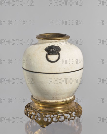 Gilded bronze mounted vase — lamp body; creator, Chinese; Qing dynasty 1644–1911; decorative vessel/vase used as a lamp body; cracklework decoration; 18th century; China; height 33 cm; provenance unknown., A rounded ceramic vessel in off-white with a crackled surface glaze sits on an ornate metallic stand. The vessel has a narrow neck with a metallic rim in a warm golden color and a dark decorative ring handle attached to a sculpted dark motif on the body. A thin dark band encircles the vessel near its midsection. The stand beneath is an intricate openwork design in a golden color with curving, scroll-like elements and a reflective surface beneath. The visible colors include off-white, dark brown or black, and gold.