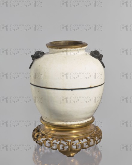 Gilded bronze mounted vase-lamp body, maker, Chinese; Qing dynasty 1644–1911; decorative vessel; vase/lamp with cracklework decoration; 18th century; China; vessel; height 33 cm., A rounded ceramic vase with a pale off-white glaze and a subtle crackle texture sits on a decorative metallic stand; the vase has a short neck with a brass-colored rim and a faint dark horizontal line around its midsection, and a pair of small dark sculpted handles near the shoulder that resemble stylized animal heads. The stand is ornamental and metallic with a warm gold tone and an openwork scrolling pattern, and the overall color palette includes off-white, dark brown to black accents on the handles and rim, and gold on the rim and stand.