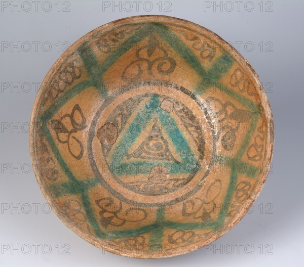 Bowl, Islamic art, Arabic culture, ceramic bowl with geometric ornament; decoration on the inner grid: geometric-spiral motif; 12th–15th century; North Africa; vessel; weight 0.72 kg; height 10 cm; diameter 21.8 cm, A shallow circular ceramic bowl viewed from above with a worn, earthy tan surface decorated with geometric and curvilinear patterns in green and dark brown, including a central triangular motif surrounded by concentric circular bands and a ring of alternating triangular and petal-like shapes; the glaze shows areas of wear and a few small chips exposing rougher clay, and the background behind the bowl is a smooth pale gray.