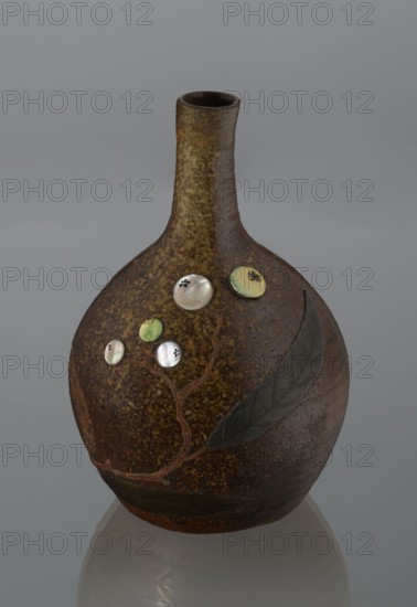 Sake bottle tokkuri, gift; Japanese, Bizen, 19th century Edo–Meiji. A bottle with a spherical belly and a narrow cylindrical neck; covered with brown glaze that becomes light coffee-coloured and spotted on the upper belly and neck. Belly decorated with a branch of biwa Japanese medlar, fruits in green-yellow glaze inlaid with mother-of-pearl, leaves made using sprinkling on black lacquer. Vessel; height 18 cm, diameter 11.8 cm., A rounded ceramic vase with a narrow neck sits against a plain grey background, showing a mottled brown and ochre surface with darker brown areas and a slightly glossy finish; a raised vine-like branch in reddish-brown winds across the body connecting a few circular decorative inlays that appear pearlescent white and pale green, and a pair of large dark grey leaves in relief are attached to the lower side of the vessel.