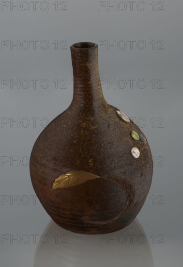Sake bottle tokkuri, Japanese, Bizen; 19th century Edo/Meiji. A bottle with a spherical belly and a narrow cylindrical neck; covered with brown glaze that in the upper belly and on the neck turns light coffee-colored and becomes spotted. Decorated on the belly with a branch of biwa Japanese medlar, fruits made with green-yellow glaze and inlaid with mother-of-pearl, leaves made using the technique of sprinkling on black lacquer. Vessel; height 18 cm, diameter 11.8 cm., A rounded ceramic vessel with a narrow neck sits against a neutral gray background, showing a textured surface in shades of brown and dark brown with speckled lighter brown areas; a metallic gold leaf-shaped ornament is applied to the body, and a few iridescent circular discs in pale pink, green, and pearly tones are attached near the upper curve, with the bottom reflecting softly on the surface below.