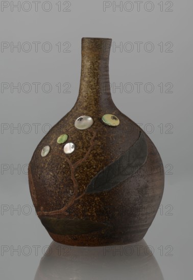 Sake bottle tokkuri, noted. Japanese, Edo–Meiji period 19th century. Bizen ware. A bottle with a spherical belly and a narrow cylindrical neck; covered with brown glaze that in the upper belly and on the neck turns light coffee-colored and becomes spotted. Decorated on the belly with a branch of biwa Japanese medlar, fruits made with green-yellow glaze and inlaid with mother-of-pearl, leaves made using a sprinkling technique on black lacquer. Vessel; height 18 cm, diameter 11.8 cm., A rounded ceramic vase with a narrow neck sits against a neutral gray background. The vase surface is mottled brown with darker and lighter areas, and it features raised decorative elements including a few leaf shapes in dark green and olive tones and a winding branch in a reddish-brown. Several circular, glossy embellishments in pearly white and pale green are set along the branch, some showing subtle iridescence. The neck of the vase is a lighter, speckled brown while the lower body shows smoother, darker...