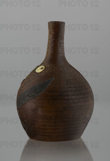 Sake bottle tokkuri, Japanese, Bizen workshop, Edo–Meiji period 19th century. Stoneware bottle with spherical belly and narrow cylindrical neck, height 18 cm, diameter 11.8 cm. Covered with brown glaze that becomes light coffee-colored and spotted on the upper belly and neck. Decorated on the belly with a branch of biwa Japanese medlar: fruits in green-yellow glaze inlaid with mother-of-pearl and leaves made using sprinkling on black lacquer. Function: vessel for sake., A rounded ceramic vessel with a narrow neck and small opening sits upright against a neutral background; the vessel has a textured surface in various shades of brown with subtle darker and lighter brown banding, a matte finish, and visible surface irregularities; a dark gray leaf-shaped applique rests on one side, and a pale circular decorative disk with a darker center is positioned nearby; the vessel casts a soft reflection on the smooth surface beneath it, and the background appears in muted gray tones.
