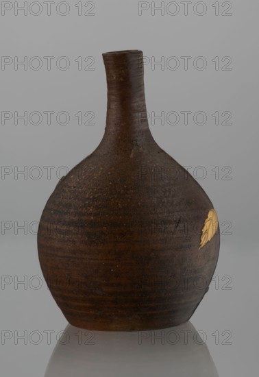 Sake bottle tokkuri, maker, Japanese, Bizen; 19th century Edo–Meiji. Ceramic vessel with spherical belly and narrow cylindrical neck; height 18 cm, diameter 11.8 cm. Covered with brown glaze that becomes light coffee-colored and spotted on the upper belly and neck. Belly decorated with a branch of biwa Japanese medlar: fruits in green-yellow glaze inlaid with mother-of-pearl, leaves made using sprinkling on black lacquer., A rounded ceramic vessel with a narrow, slightly curved neck sits against a plain light gray background, showing a textured surface in dark brown and reddish-brown tones with subtle horizontal striations and areas of darker mottling; a small golden-yellow leaf-shaped decoration is visible on one side, and the vessel rests on a reflective surface that mirrors its base faintly.