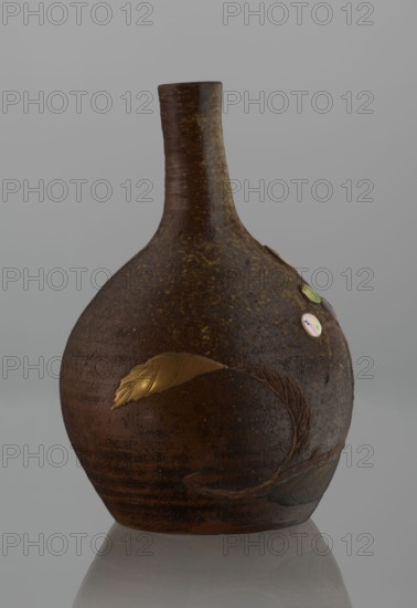 Sake bottle tokkuri, Japanese, Bizen; 19th century Edo–Meiji. Stoneware vessel with spherical belly and narrow cylindrical neck, height 18 cm, diameter 11.8 cm. Covered with brown glaze that lightens to a coffee color and becomes spotted on the upper belly and neck. Belly decorated with a branch of biwa Japanese medlar: fruits in green-yellow glaze inlaid with mother-of-pearl, leaves executed by sprinkling on black lacquer., A rounded ceramic vessel with a narrow neck sits against a plain gray background and reflected surface; the vessel has a rough, mottled brown surface with darker and lighter brown variations and speckled texture, a few glossy iridescent round inlays in soft green and pink tones near the upper side, and decorative raised leaf shapes in metallic gold and muted gray set into the body; a curved raised vine-like line wraps around the lower side, and the neck shows horizontal wheel-thrown ridges in brown.