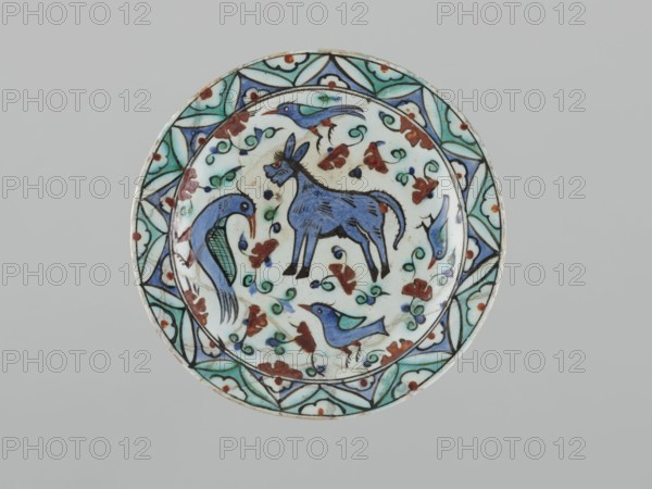 Plate, Islamic art, Turkish, Ottoman dynasty 1299–1922, ceramics, Iznik, 17th century. Dish vessel, height 5.3 cm, diameter 26.5 cm, weight 0.62 kg. Decoration: in the mirror a donkey, four birds and a floral ornament; on the rim geometric-floral; stylized flowers, animals, birds, donkeys; geometric and floral ornament., A circular ceramic plate with a decorative rim and central painted scene, predominantly showing colors of white, blue, turquoise, green, brown, and black; the rim features repeating geometric and floral motifs in turquoise, green, blue, and brown outlined in black against a white background; the central area depicts a blue animal with black outlines and markings standing amid swirling vine-like green tendrils and small brown floral shapes, accompanied by several stylized birds in shades of blue and turquoise with black details; the overall background of the plate is white with scattered decorative accents and a thin black outline near the inner edge.