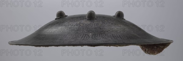 Shield sipar, Persian, Qajar dynasty 1785–1925, Iranian, 18th/19th century. Material: metal weapon; height 9.0 cm, diameter 47.5 cm. Decoration: field with hunting scenes hunters, horses, archers, belts ornamented with plant tendrils and inscriptions stylized in Kufic script. Function: militaria., A domed metal object with a low, wide profile and a scalloped rim, shown against a pale gray background, featuring a dark metallic gray surface with intricate incised decorative patterns and a few raised rounded bosses along the top; the underside edge reveals a layer of fibrous material in muted brown and beige tones.