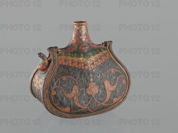 Waterskin matara, Islamic/Turkish, Ottoman dynasty 1299–1922, 2nd half of the 16th century, Turkey. Kidney-shaped leather waterskin with an appliqué of a colorful lizard; bottle with rounded bottom, narrow neck for pouring and small spout for drinking. Decorated with applied arabesques cut from shagreen and arabesques painted with gold. One narrow side bears a protective emblem in the form of a six-pointed star Seal of Solomon intended to ensure the water's freshness. The leather matara is a copy of gold bottles set with precious stones used at the sultan's court and may have been used during military expeditions. Dimensions: height 23.0 cm, width 24.5 cm, depth 14.0 cm, weight 0.62 kg., An ornately decorated curved container with a narrow neck and a short spout, shown against a plain light gray background, featuring a textured surface with swirling vine and floral motifs; the main visible colors are dark brown and black on the body, with prominent accents of red, green, and gold throu...