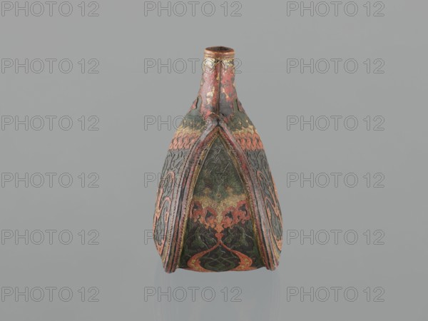 Waterskin matara, Islamic/Turkish, Ottoman dynasty 1299–1922. Kidney-shaped leather waterskin with appliqué of a colorful lizard; rounded bottom, narrow neck for pouring and small spout for drinking. Decorated with applied shagreen arabesques and arabesques painted in gold. One narrow side bears a protective six-pointed star Seal of Solomon intended to ensure the water's freshness. The leather matara is a copy of gold, gem-set court bottles; it may have been used by the sultan on military expeditions. Unique type — four such mataras are known worldwide. 2nd half of the 16th century, Turkey. Vessel; height 23.0 cm, width 24.5 cm, depth 14.0 cm, weight 0.62 kg., An ornate, narrow-necked vessel stands upright against a plain gray background; the vessel displays a symmetrical, elongated teardrop shape with a pointed top and a wider base. The surface shows layered, decorative panels with swirling, curvilinear patterns and scalloped edges. Visible colors include muted red, deep green, dark g...