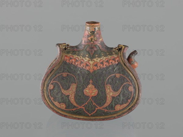 Waterskin matara, Turkish, Ottoman dynasty 1299–1922, 2nd half of the 16th century. Kidney-shaped leather waterskin with an appliqué of a colorful lizard; bottle with rounded bottom, narrow neck for pouring and a small spout for drinking. Decorated with applied arabesques cut from shagreen and arabesques painted with gold. One narrow side bears a protective six-pointed star emblem Seal of Solomon intended to ensure the water's freshness. The leather matara is a copy of gold bottles set with precious stones used at the sultan's court; such leather bottles may have been used by the sultan during military expeditions. Only four comparable mataras are known worldwide. Vessel waterskin — height 23.0 cm, width 24.5 cm, depth 14.0 cm, weight 0.62 kg., An ornately decorated flat flask-shaped object with a rounded base and a short cylindrical neck, featuring a curved spout on one side and a small broken area on the rim, shown against a plain light gray background; the surface displays intricate...