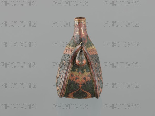Waterskin matara, Ottoman Turkey, 2nd half of the 16th century. Kidney-shaped leather waterskin with rounded bottom, narrow neck and small spout for drinking; height 23.0 cm, width 24.5 cm, depth 14.0 cm, weight 0.62 kg. Decorated with applied shagreen arabesques and arabesques painted in gold; one narrow side bears a protective emblem in the form of a six-pointed star Solomon’s Seal intended to ensure the water's freshness. The leather matara is a copy of gold bottles set with precious stones used at the sultan's court and may have been used during military expeditions. Only four comparable leather mataras are known worldwide., A decorative vessel with a tall narrow neck and a rounded body is shown against a plain gray background; the surface of the vessel displays ornate carved and painted patterns in a palette of green, red, orange, gold, and black with areas of worn patina, and the front features a raised tear-shaped motif and symmetrical flared panels with intricate swirling desig...
