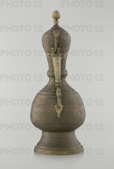 Jug aftabe, aftabe, geometric and plant ornament, dragons, zoomorphism. Decoration: dense plant and geometric ornament. Persian, Qajar dynasty 1785–1925, 19th century. Materials: vessel. Place of origin: Iran possibly Central Asia. Dimensions: height 34.0 cm; width 22.7 cm; diameter 13.3 cm., An ornate metal vessel with a tall, elongated silhouette sits against a plain light gray background, featuring a bulbous lower body that tapers into a narrow neck and rises to a domed lid topped with a finial; the surface shows intricate raised and incised patterns in varying shades of dark bronze and muted gold, with areas of reddish-brown patina and pale greenish tones on decorative metal fittings; a long vertical strap-like handle runs from the lid to the lower body, embellished with small textured motifs and a pointed terminal piece; the base flares outward and is decorated with repeating geometric and floral banding, and the whole object rests on a reflective surface that casts a faint mirror...