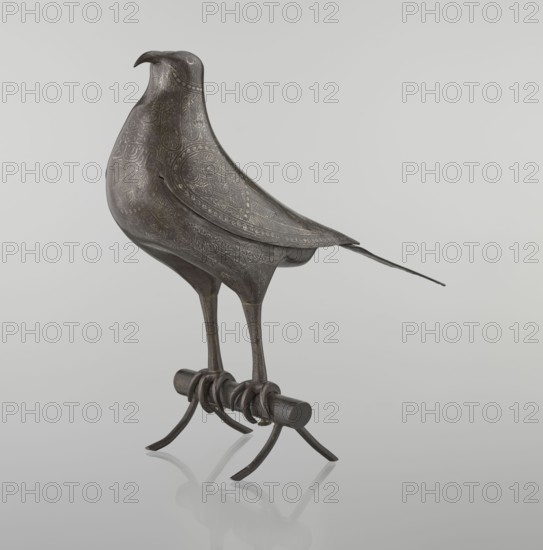 Falcon on a branch, Islamic Persian, 18th century, Iran; sculpture/figurine of a bird falcon with feathers, inscriptions/calligraphy; figurine, height 28.5 cm, width 27.5 cm, depth 14.5 cm, weight 1.25 kg., A sculpted bird figure rendered in dark brown metal with etched decorative patterns across its body and wings, standing on slender legs attached to a short cylindrical perch with curled metal supports and curved feet, set against a smooth light gray background with a faint reflection beneath the perch.