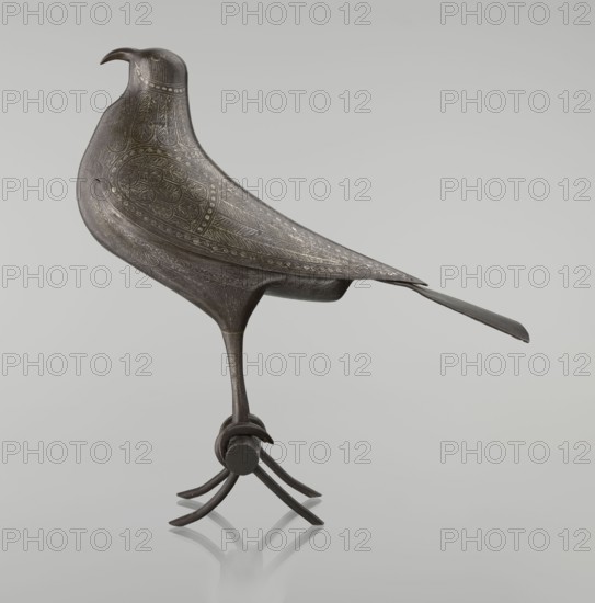 Falcon on a branch, Islamic art, Persian culture, sculpture/figurine of a bird falcon, feathers, inscriptions/calligraphy, 18th century, Iran, figurine, height 28.5 cm, width 27.5 cm, depth 14.5 cm, weight 1.25 kg, A sculpted bird figure in dark metallic gray with lighter grayish decorative etched patterns across its body and wings stands on a single slender leg attached to a small cylindrical base and thin curved feet, with a long tapered tail extending behind it; the surface shows intricate feather-like engravings and a smooth curved beak, and the scene is set against a pale neutral gray background.
