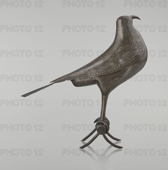 Falcon on a branch, Islamic art, Persian culture, sculpture/figurine of a bird falcon, feathers, inscriptions, calligraphy, 18th century, Iran, figurine; height 28.5 cm, width 27.5 cm, depth 14.5 cm, weight 1.25 kg, A sculpted bird form with a smooth, dark metallic surface featuring engraved feather patterns and decorative motifs, standing on a slender leg that connects to a circular joint and curved talons, with a long tapered tail extending backward; the bird has a rounded head with a small hooked beak and subtle surface texture; the overall colors visible are dark gray, black, and hints of brown against a pale gray background.