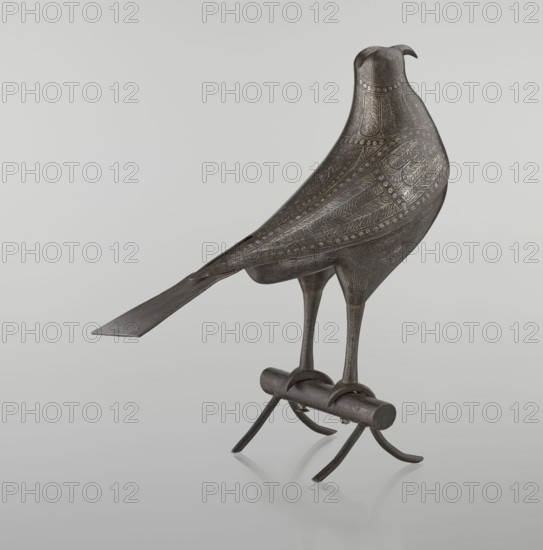 Falcon on a branch, Islamic Persian, 18th century Iran, figurine sculpture of a bird falcon with feathers, inscriptions/calligraphy; height 28.5 cm, width 27.5 cm, depth 14.5 cm, weight 1.25 kg., A sculpted bird figure made of dark metal with engraved decorative patterns covering its body and wings, perched on a short cylindrical perch with curved supporting legs; the bird has a long, flat tail, slender legs, clawed feet gripping the perch, and a slightly curved beak. The visible colors are dark gray, muted bronze, and subtle lighter highlights, set against a pale gray background.