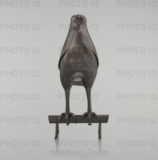 Falcon on a branch, Islamic art, Persian culture, sculpture/figurine of a bird falcon, feathers, inscriptions, calligraphy, 18th century, Iran, figurine; height 28.5 cm, width 27.5 cm, depth 14.5 cm, weight 1.25 kg, A dark bronze bird-shaped object with a smooth rounded body and stylized head stands on long slender legs that end in curved talons gripping a horizontal perch; the surface of the bird shows intricate incised patterns and faint areas of lighter patina, and the scene includes shades of dark brown, gray, and hints of greenish bronze against a light gray background.