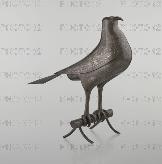 Falcon on a branch, Persian, 18th century, Iran; sculptural figurine of a bird falcon with feathers, inscriptions/calligraphy; material unspecified; figurine height 28.5 cm, width 27.5 cm, depth 14.5 cm, weight 1.25 kg., A sculpted bird-shaped object in dark metallic gray stands on a short cylindrical perch with curved claw-like supports; the bird has a long tail and slender legs, with finely etched decorative patterns across its body and wings, and a smooth, slightly reflective surface showing hints of lighter gray where light catches it, against a pale gray background.