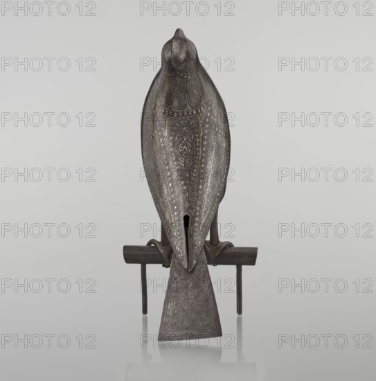 Falcon on a branch, 18th century, Iran; Persian Islamic sculpture/figurine of a falcon with feathers, inscriptions/calligraphy; figurine; height 28.5 cm, width 27.5 cm, depth 14.5 cm, weight 1.25 kg., A sculpted bird figure in dark gray tones sits on a simple horizontal perch with two vertical supports, the bird shown from the back with a tapered tail extending down and resting between the supports; the bird's surface is covered in intricate engraved patterns and textures forming symmetrical feather-like motifs in lighter gray, giving a subtle contrast against the darker base color, and the perch appears in dark brownish-gray with a slight reflective shadow on the light gray background.