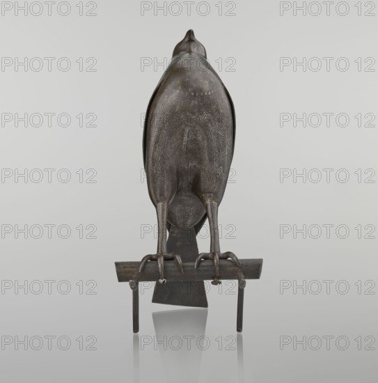Falcon on a branch, Persian, 18th century, Iran; sculpture/figurine of a bird falcon with feathers, inscriptions/calligraphy; figurine; height 28.5 cm, width 27.5 cm, depth 14.5 cm, weight 1.25 kg., A sculpted bird figure viewed from behind, perched on a horizontal bar supported by vertical posts, with carved detailing on its back and wings; the bird and stand have a dark metallic brown-gray surface with subtle lighter highlights and a few areas of patina that appear greenish and yellowish, set against a smooth pale gray background with a soft reflection beneath the stand.