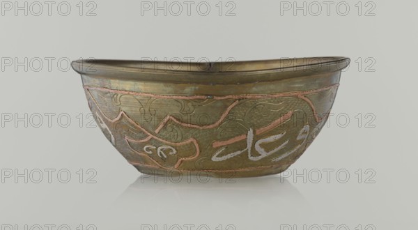 Bowl, Islamic art, Arabic culture, arabesque ornament, calligraphy, Arabic inscriptions, geometric motifs, 19th century, Middle East, possibly Syria, vessel; height 4.9 cm, diameter 13.3 cm, A shallow metal bowl with a rounded rim sits against a pale gray background, showing a metallic surface in shades of muted gold and brass with areas of darker patina. The bowl's exterior is decorated with raised sinuous designs in a coppery pink tone and lighter inlaid strokes in an off-white shade, set against a textured background of small, dimpled indentations. The rim and base edges appear slightly darker olive-brass, and the overall finish shows subtle wear and variations in color from greenish-bronze to warm brown. The bowl rests on a reflective pale surface that produces a soft, blurred mirror image beneath it.