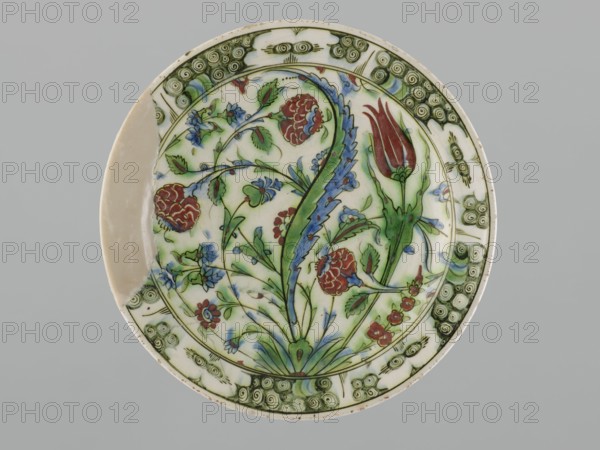 Plate;, Earthenware vessel Iznik, Turkey, 17th century; height 5.5 cm, diameter 30.5 cm. Decoration: hyacinths, roses, tulips, saz motifs, rim spirals., A round ceramic plate with a decorative floral design featuring a large tulip-like bud and several stylized flowers and leaves in shades of green, red, blue, and pale blue on a white background, surrounded by a green patterned border with spiral motifs; a section of the plate shows an unglazed beige area where a piece is missing, and the overall scene is set against a neutral gray background.