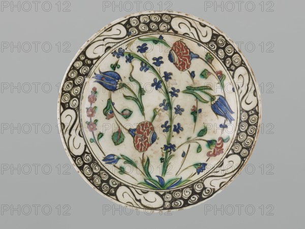 Plate, gift, Islamic art, Turkish, Ottoman dynasty 1299–1922, ceramics decorated with flowers carnations, hyacinths, roses, tulips, rim decoration: spirals, 16th/17th century, Iznik, Turkey; vessel, height 6.3 cm, diameter 29.2 cm, A round ceramic plate with a central floral design surrounded by a decorative border; the plate shows a cream or off-white background with painted flowers and stems in blue, green, and red, with touches of pink and dark green leaves; the floral motifs include tulip-like and carnation-like shapes and small clustered blossoms, all depicted with thin green stems and occasional blue buds; the border features a repeating scroll and spiral pattern in black and cream, and the plate edge shows slight wear and a thin brown rim; the overall color palette includes cream, blue, green, red, pink, black, and brown.