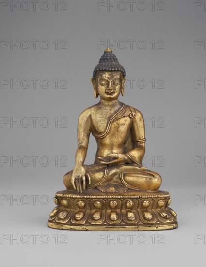 Shakyamuni Buddha, buddha iconographic, lotus throne iconographic, Himalayan culture, Tibetan Buddhist art, Buddha ca. 560–480 BCE, ushnisha, mudras, 19th century, Tibet, sculpture; height 10.2 cm, width 7.3 cm, depth 5.4 cm, A gilded statue of a seated figure on a decorative pedestal, showing a golden body with a darker blue-black textured hairpiece and a small golden point at the top, the figure seated cross-legged with one hand resting on the knee and the other held near the lap, wearing a draped garment over one shoulder with visible folds and ornamental details, the pedestal featuring a repeating rounded motif and a golden finish, all set against a plain gray background with neutral tones.