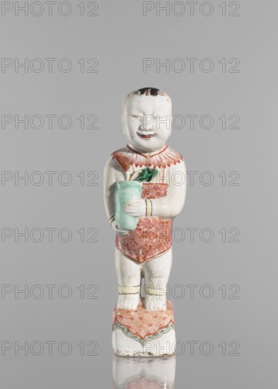 One of the Hehe Er Xian brothers holding a vase with lotus buds;, Chinese; Qing dynasty, Kangxi period 1662–1722; colored porcelain; subject includes lotuses, flower vase, children/boys; sculpture; height 28.0 cm; weight 0.75 kg., A ceramic figurine of a child standing on a decorated rectangular base, with a pale white glaze overall and painted details in red, green, black, and a pale yellow; the child has a rounded face with closed eyes, a small smiling mouth, and a black painted patch on the top of the head, wearing a red patterned garment with a ruffled red collar and pale yellow bands at the wrists and ankles; the child holds a pale green vase containing a red flower and green leaves, and the base shows red patterned decoration with green and black scalloped edging against the white ceramic.