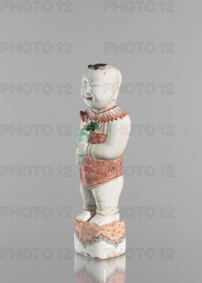 One of the Hehe Er Xian brothers holding a vase with lotus buds; maker, Chinese; Qing dynasty, Kangxi period 1662–1722; colored porcelain; subject includes lotuses, flower vase, children/boys; sculpture; height 28.0 cm; weight 0.75 kg; date range 1662–1722; China., A ceramic figurine of a childlike figure standing on a small decorated pedestal, with a pale white body, black hair detail, and painted facial features; the figure wears a patterned red garment and red collar detail, and holds a green object close to the chest with a small brown element atop it; the pedestal and garment have additional green and red decorative patterns, and there are subtle beige and brown aged marks on the base.