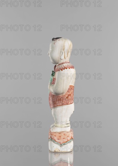 One of the Hehe Er Xian brothers holding a vase with lotus buds, Chinese, Qing dynasty, Kangxi period/style 1662–1722, colored porcelain, lotuses, flower vase, children/boys, sculpture, height 28.0 cm, weight 0.75 kg, dated 1662–1722, China., A glazed ceramic figurine of a standing child shown in profile against a neutral gray background, with a pale off-white skin tone and fine painted hair details in dark brown and light brown on the head; the child wears a short sleeved garment with a patterned sash in reddish orange and a collar with reddish orange feather-like decoration, and holds a small green object near the chest; the lower garment and pedestal are painted with reddish orange textured patterns and pale green decorative borders, with subtle yellowing and darker spots on the glaze visible in places.