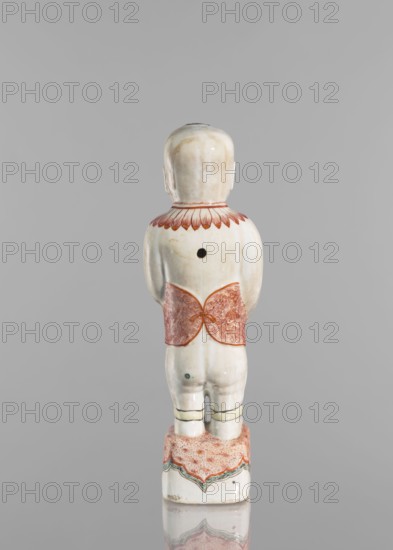One of the Hehe Er Xian brothers holding a vase with lotus buds; creator, Chinese; Qing dynasty, Kangxi period 1662–1722; colored porcelain; subject includes lotuses, flower vase, children/boys; sculpture; height 28.0 cm; weight 0.75 kg., A ceramic figurine shown from the back standing on a decorated rectangular base, with a glossy off-white surface and areas of light beige shading; the figure wears a red patterned wrap around the waist and a red scalloped collar design at the shoulders, and there are additional decorative bands in green and red on the base and near the lower legs; the head is rounded and smooth, the arms rest at the sides, and a dark circular hole is visible on the upper back; colors visible include off-white, beige, red, green, and black.