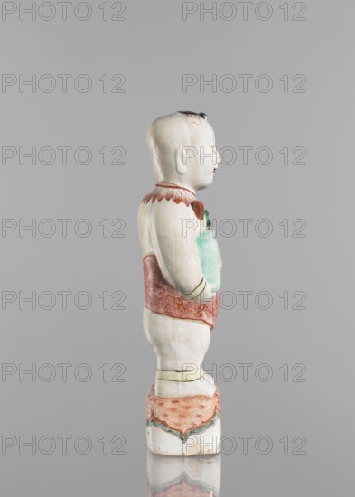 One of the Hehe Er Xian brothers holding a vase with lotus buds;, Chinese; Qing dynasty, Kangxi period 1662–1722; colored porcelain; subject includes lotuses, flower vase, children/boys; sculpture; height 28.0 cm; weight 0.75 kg; dated 1662–1722., A ceramic figurine of a child seen in profile standing on a decorated base, with a pale white body and subtle surface variations, wearing a red patterned garment around the waist and a red decorative collar, holding a pale green object against the chest, and with small touches of black on the hair and facial features; the base and garment include accents of green and the overall background is a uniform light gray.