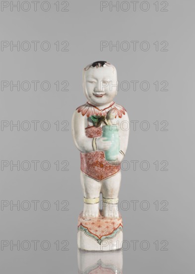 One of the Hehe Er Xian brothers holding a vase with lotus buds, Chinese, Qing dynasty, Kangxi period 1662–1722, colored porcelain sculpture depicting boys/children with lotuses, height 28.0 cm, weight 0.75 kg., A ceramic figurine of a child standing on a decorated base, painted primarily in white with accents of red, green, black, yellow, and pale turquoise; the child has a rounded face with closed eyes and a small smile, short black hair painted on the top of the head, a red patterned collar and red patterned garment around the waist, pale yellow bands around the lower legs, and is holding a pale turquoise vessel with a green leaf and a small beige object emerging from it; the base is white with a red patterned top surface and a scalloped green and red decorative border.