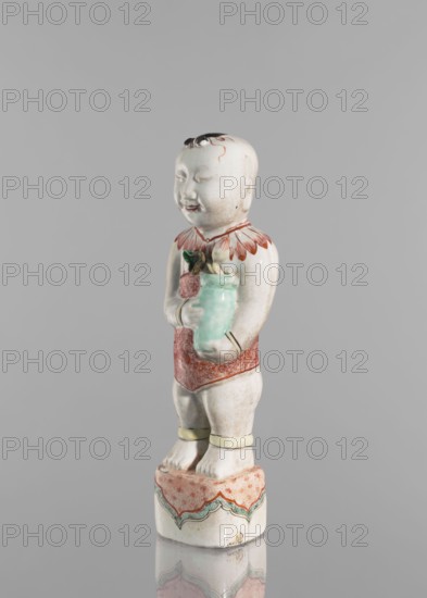 One of the Hehe Er Xian brothers holding a vase with lotus buds;, Chinese, Qing dynasty, Kangxi period 1662–1722; colored porcelain sculpture; subject: boys/children and lotuses; height 28.0 cm; weight 0.75 kg; dated 1662–1722; China., A ceramic figurine of a standing child on a decorated pedestal, shown against a plain gray background. The child has a pale off-white complexion with subtle shading, short dark hair painted on top of the head, and a smiling facial expression with closed eyes. The figurine wears a red ornamental collar with petal-like shapes and a red patterned loincloth, and pale yellow anklets around the lower legs. The child holds a glossy light green vase with a few green leaves and a darker element at the top. The pedestal is mainly off-white with a pinkish-red patterned top and scalloped green and red decorative bands along the sides. Colors visible include off-white, gray, red, pinkish-red, green, light green, dark green, yellow, and black.