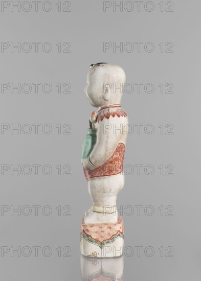 One of the Hehe Er Xian brothers holding a vase with lotus buds; maker, Chinese porcelain sculpture; colored porcelain; Qing dynasty, Kangxi period 1662–1722, China; subject: boys/children and lotuses; height 28.0 cm; weight 0.75 kg; dated 1662–1722., A glazed ceramic figurine of a child shown in profile on a square base, predominantly white with areas of pale green and reddish-orange decoration, the child wearing a patterned reddish-orange garment around the midsection and a reddish-orange collar with petal-like shapes, the child holding a pale green object against the chest, the head smooth and white with small painted dark accents at the hairline and ear, the square base decorated with reddish-orange surface patterning and a scalloped band in green and reddish-orange, the overall background a neutral grey.