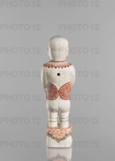 One of the Hehe Er Xian brothers holding a vase with lotus buds, Chinese, Qing dynasty Kangxi, 1662–1722, colored porcelain sculpture of boys/children with lotuses, height 28.0 cm, weight 0.75 kg, dated 1662–1722., A small pale figurine viewed from behind standing on a rectangular base, with a smooth rounded head and visible ears, a short neck rimmed by petal-like decoration in reddish tones, a cylindrical torso with a dark circular hole on the upper back, a decorative reddish patterned band wrapped around the lower torso, short legs with pale yellow bands near the knees, and a base decorated with a mottled reddish surface and scalloped green and reddish edging; visible colors include pale off-white, reddish, yellow, green, and hints of gray.