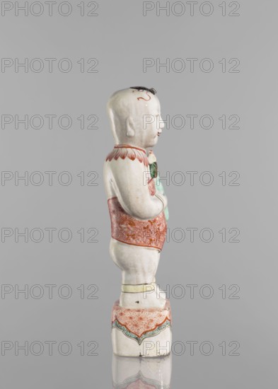 One of the Hehe Er Xian brothers holding a vase with lotus buds; maker, Chinese; Qing dynasty, Kangxi period/style 1662–1722; colored porcelain sculpture depicting boys/children with lotuses; height 28.0 cm; weight 0.75 kg., A ceramic figurine of a child standing on a decorated square base is shown in profile against a plain gray background; the child is predominantly white with painted details including a red patterned garment around the torso and hips, red scalloped decoration at the shoulders, a pale green object held near the chest, and small black and red markings on the head, while the base and trims feature red, green, and pale yellow accents.