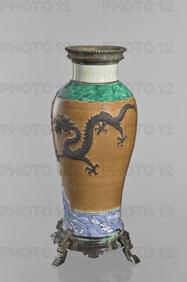 Bronze-framed vase, creator unknown, Chinese, Qing dynasty 1644–1911; decorative ceramic vessel with dragons and a pelican iconography, waves motif; plastic decoration in brown-stained biscuit; 2nd half of the 19th century, China; height 62.0 cm, width 27.0 cm., An upright ceramic vase with a rounded body and narrow neck sitting on an ornate metal stand, featuring a glossy brown main surface with a raised black dragon motif wrapping around it, a band of green glaze near the shoulder with a scalloped edge, a pale off-white neck with subtle crackle, and a blue-and-white wave-patterned lower border; the metal stand has a dark, aged finish with greenish patina.