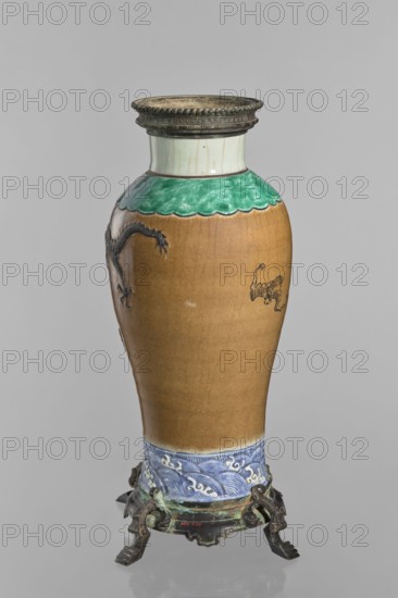 Bronze-framed vase, Chinese, Qing dynasty 1644–1911. Decorative ceramic vessel with dragons and pelican iconography, wave motifs; brown-stained biscuit plastic decoration. 2nd half of the 19th century, China. Height 62.0 cm; width 27.0 cm., A tall ceramic vase with a rounded body and narrow neck sits on an ornate metal stand; the upper rim of the vase has a metal collar in a dark metallic color with a beaded edge, followed by a pale off-white neck and a scalloped green band below it, then a warm brown main body with a few dark decorative motifs on its surface and a lower band of blue and white wave-like patterns, the metal stand beneath showing a dark, aged greenish-brown patina.