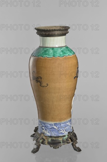 Bronze-framed vase, creator unknown, Chinese, Qing dynasty 1644–1911. Ceramic vessel with plastic brown-stained biscuit decoration depicting dragons, a pelican, waves; decorative vessel. 2nd half of the 19th century. Height 62.0 cm; width 27.0 cm., A tall, oval-shaped ceramic vase with a narrow neck and rounded shoulders sits on an ornate metal stand; the vase shows a top rim with a metal band in a dark, aged tone and a short neck glazed in pale cream with faint vertical lines, followed by a scalloped green band at the shoulder. The main body of the vase is a warm brown color with subtle surface crackling and small dark decorative swirl motifs. Near the base there is a blue and white band decorated with wave-like patterns, and the metal stand below is dark with greenish patina and has an intricate, aged design with curved legs. The visible colors include brown, green, pale cream, blue, white, dark metal tones, and hints of greenish verdigris.